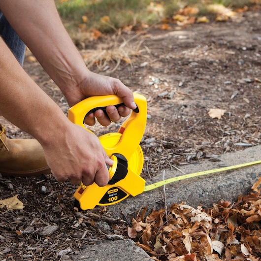 100 foot fiberglass long tape being used by a person to measure a path outdoors.